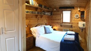 A wooden cabin bedroom with a bed, bedside tables, a window, and a shelf with books. - Astoria (Broadstairs)