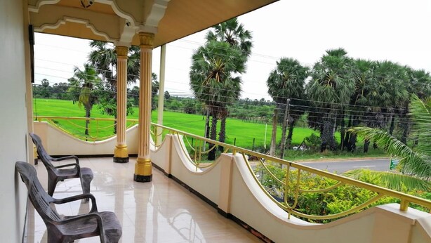 A balcony with two plastic chairs, a tiled floor, and a view of a green field with palm trees. - Victoria Guest House (Pasikuda)