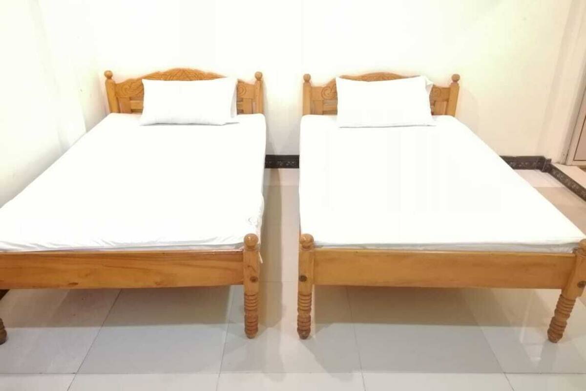 Deux lits en bois, recouverts de draps blancs, se trouvent dans une pièce aux murs blancs et au sol carrelé.