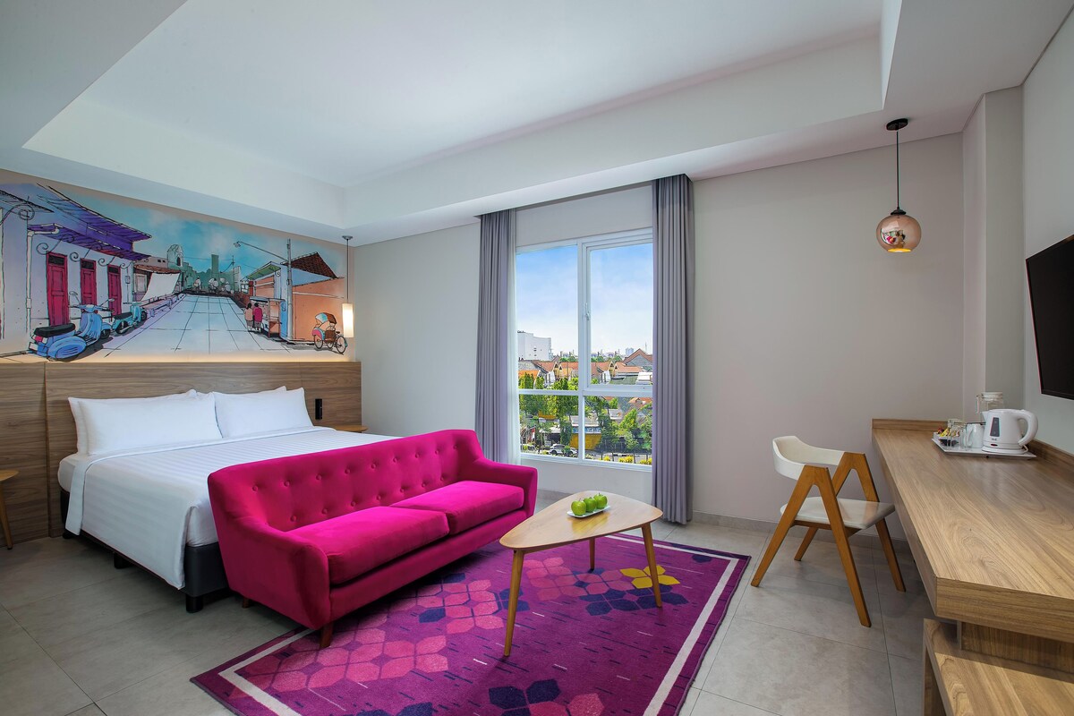 A hotel room with a bed, a pink sofa, a small table with a bowl of fruit, a wooden desk, a chair, a TV, and a large window with a city view.
