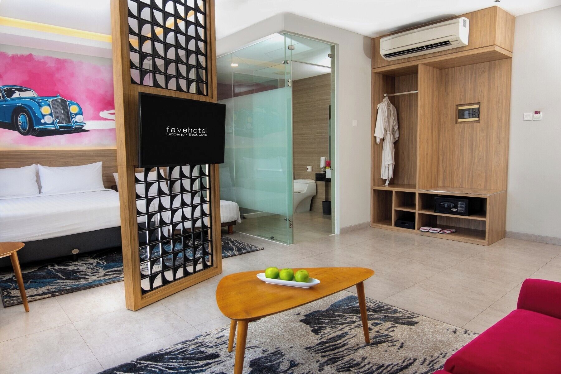 Une chambre d’hôtel avec un lit, un bureau, une chaise rouge, une armoire et une salle de bain visible à travers une porte en verre.