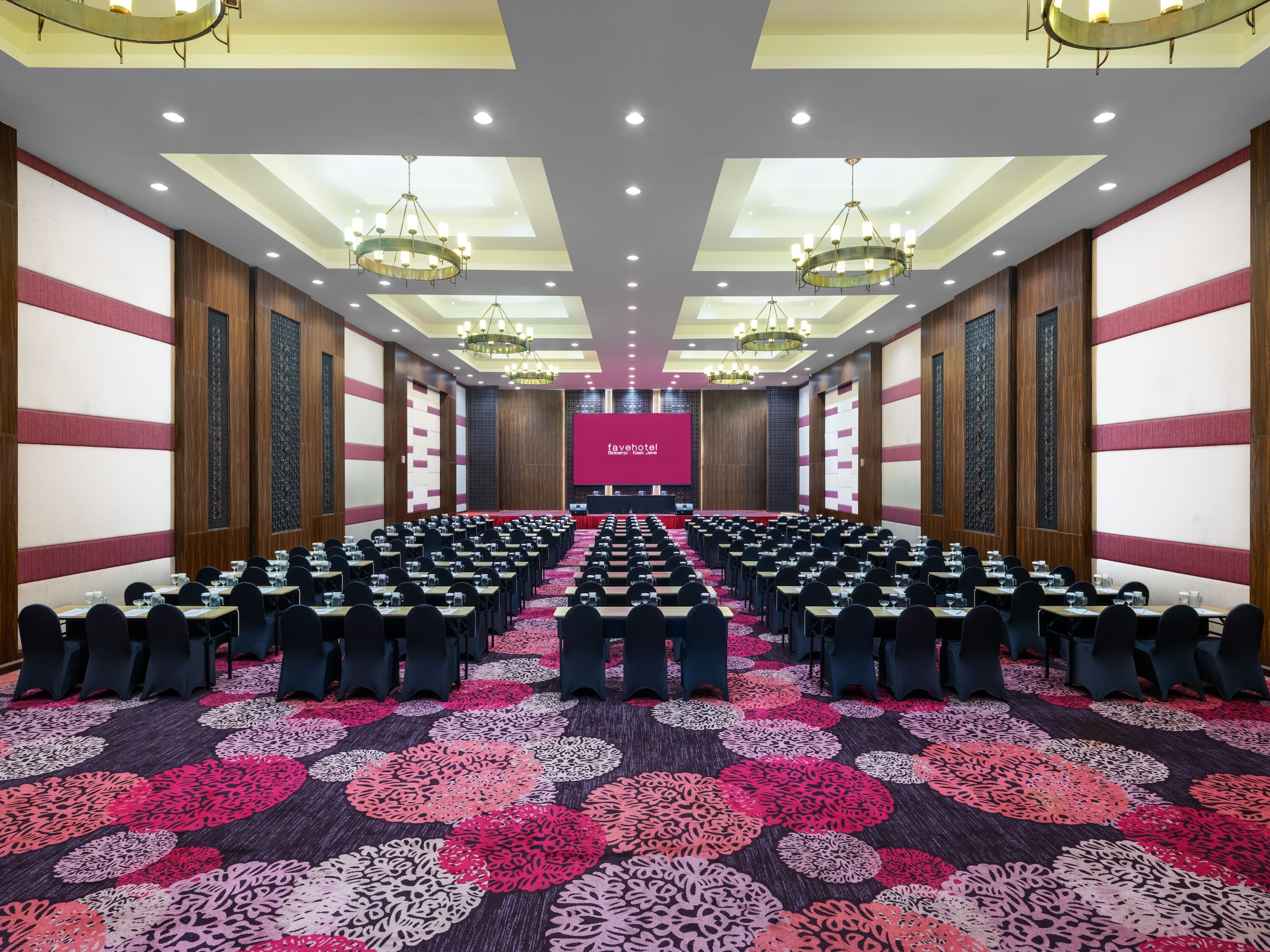 A conference room with rows of tables and chairs, a large screen at the front, and patterned carpet.