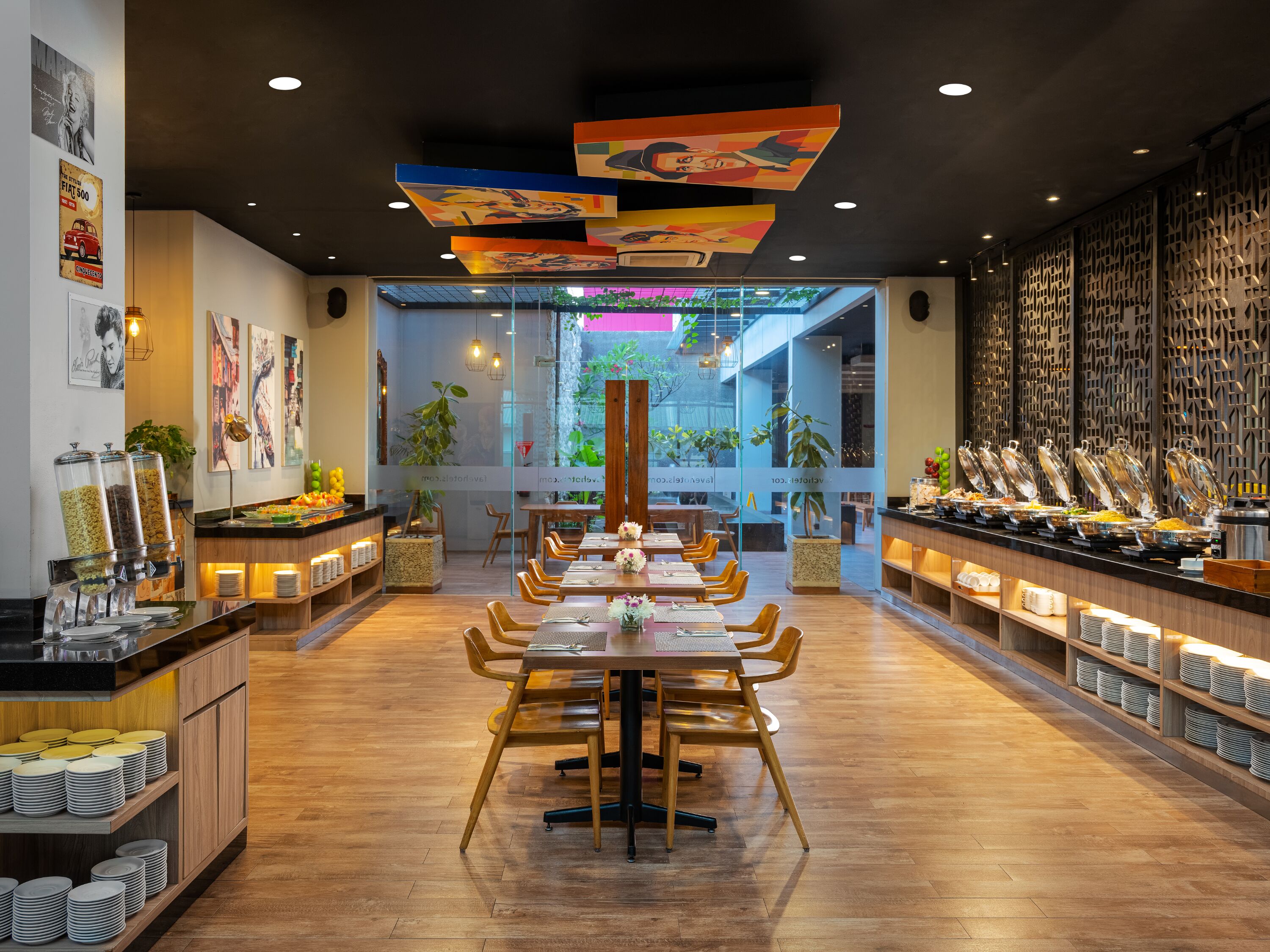 A modern restaurant with a central dining area, buffet with various food options, and a view of greenery through large windows.