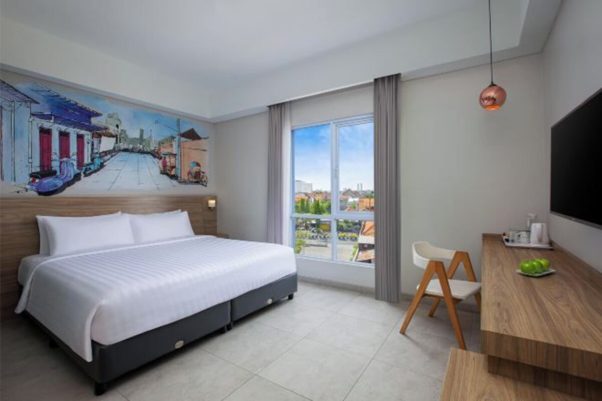 A hotel room with a bed, a desk, a chair, a painting on the wall, and a large window.