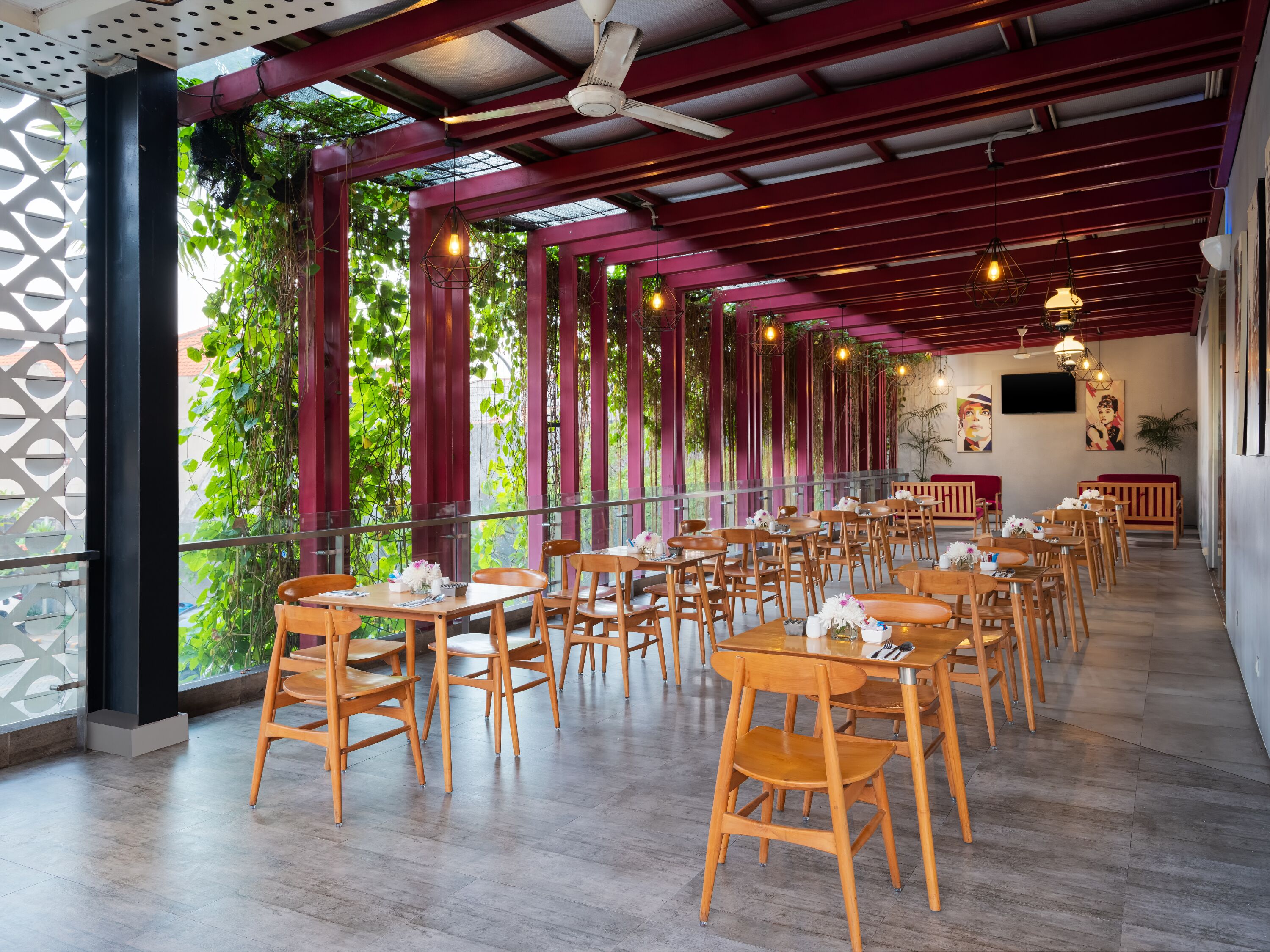 Un restaurante con mesas y sillas de madera, amplios ventanales con vegetación en el exterior y un ventilador de techo.