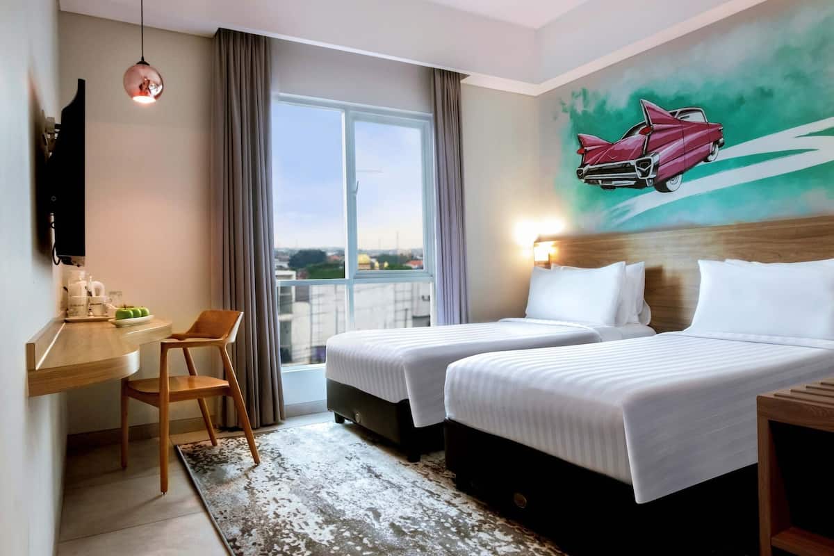Habitación de hotel con dos camas, un escritorio con silla, un televisor y un ventanal con cortinas.
