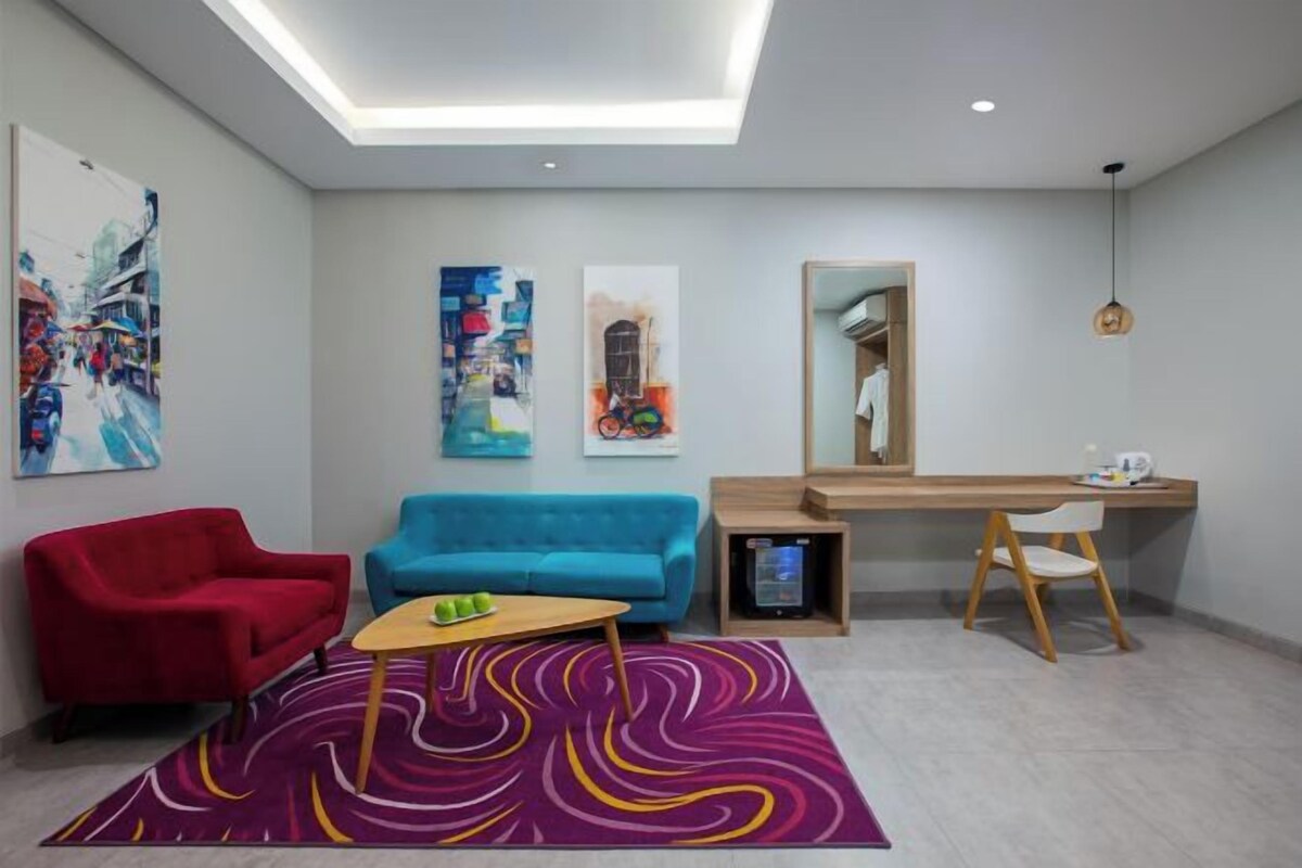A modern hotel room with a red and blue sofa, a small wooden table, a desk with a chair, and artwork on the walls.