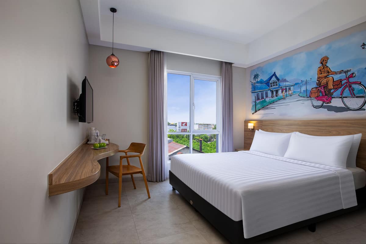Habitación de hotel con cama, escritorio con silla, un cuadro en la pared y vistas al exterior.