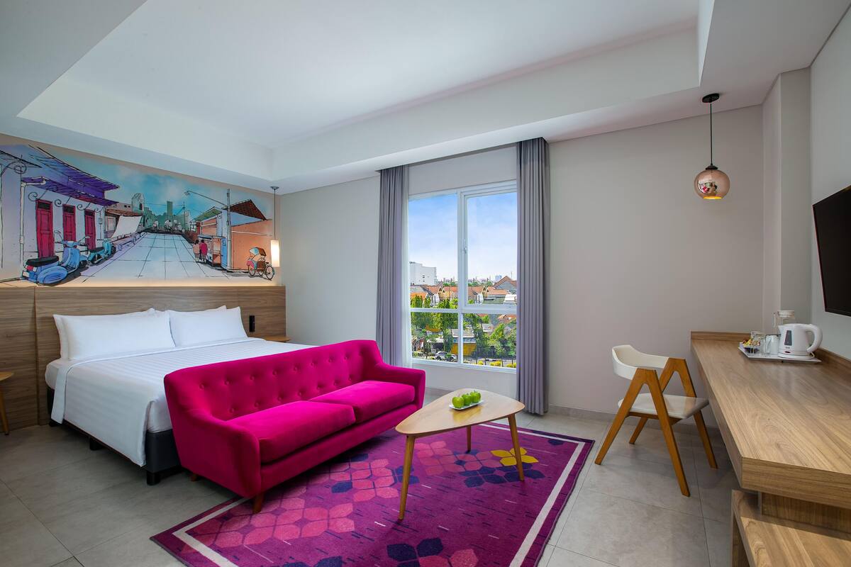 Una habitación de hotel con una cama, un sofá rosa brillante, una mesita pequeña, un escritorio de madera y un ventanal con vistas a edificios.