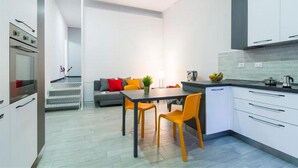A modern kitchen with white cabinets, a dining table with orange chairs, and a gray sofa in the background. - Freccia Rossa Bologna Apartment (Bologna)