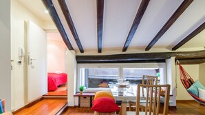 Een gezellige kamer met een houten vloer, een eettafel met stoelen, een bankje, een bed met rode beddengoed en een hangmat.