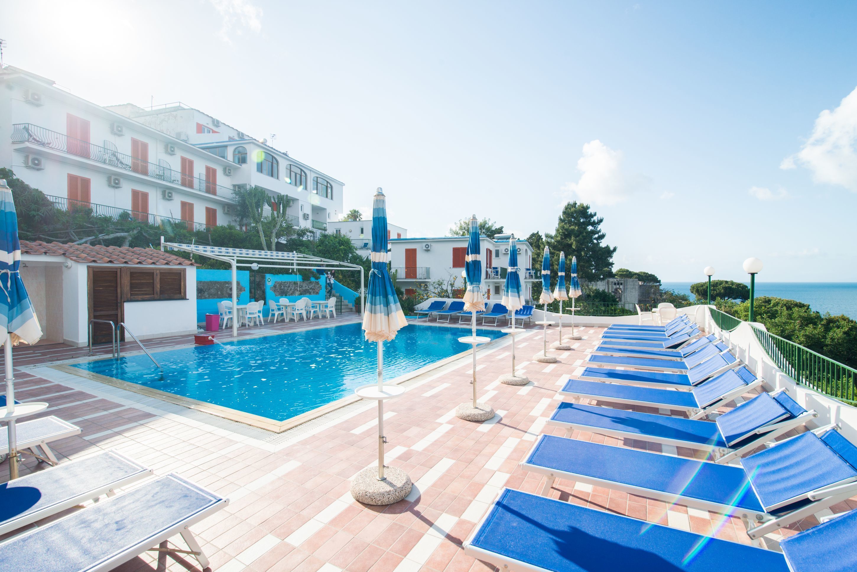 A pool area with lounge chairs and umbrellas, a clear sky, and a building with red shutters in the background.