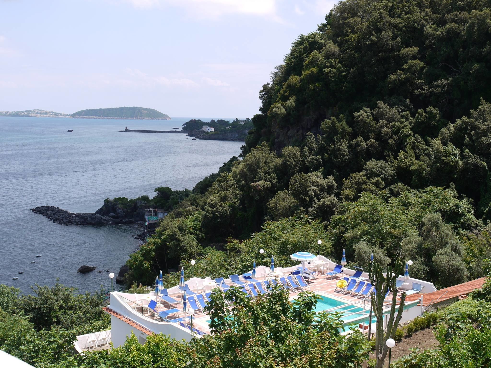 A pool area with lounge chairs and umbrellas, surrounded by lush greenery and overlooking a calm sea with a distant island.