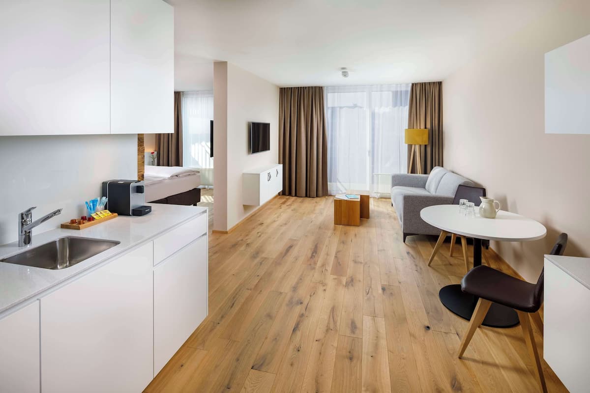 A modern hotel room with a kitchen area, a dining table, a sofa, and a large window with curtains.