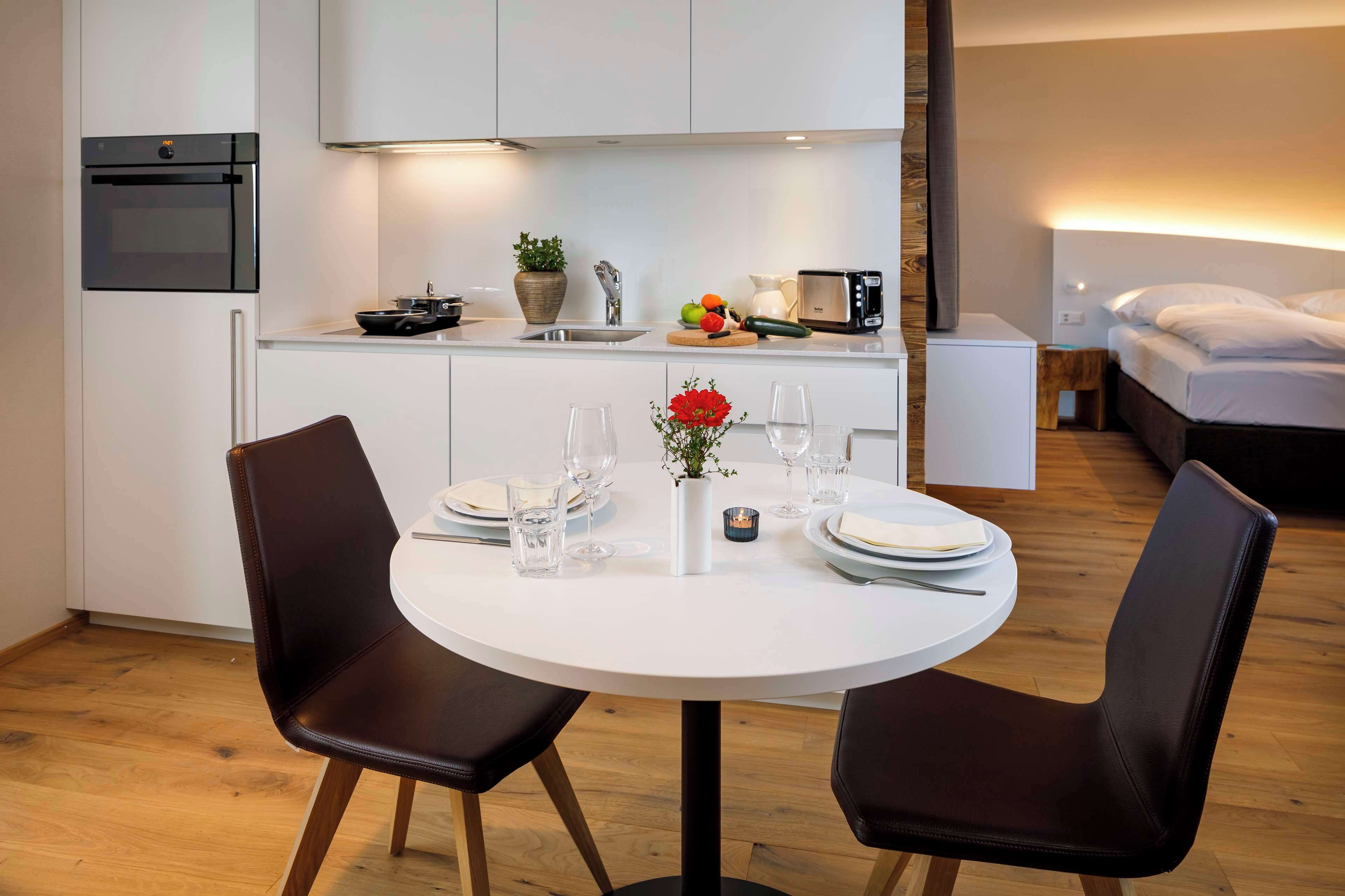 A modern kitchen with a built-in oven, a microwave, and a sink. There is a dining table with chairs, a vase with flowers, and a fruit bowl.