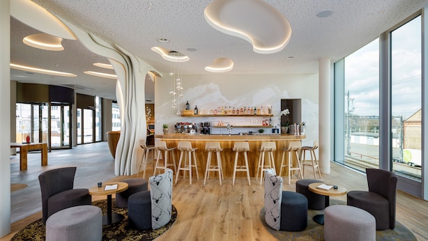 A modern bar area with a wooden bar counter, bar stools, and a seating area with round tables and chairs. - Hotel Allegra Lodge (Kloten)