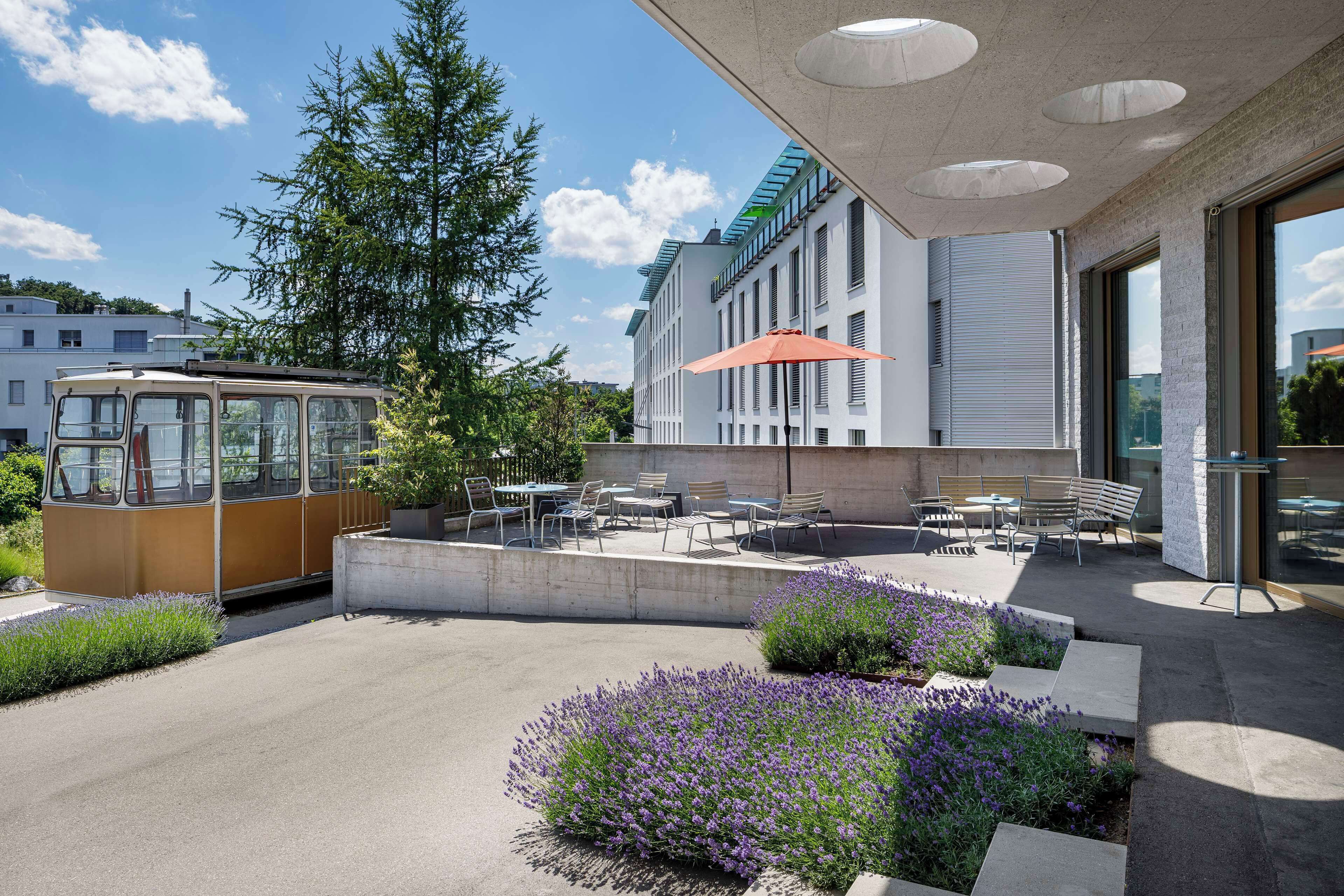 Un edificio moderno con terraza al aire libre, un tranvía y un jardín con flores moradas.