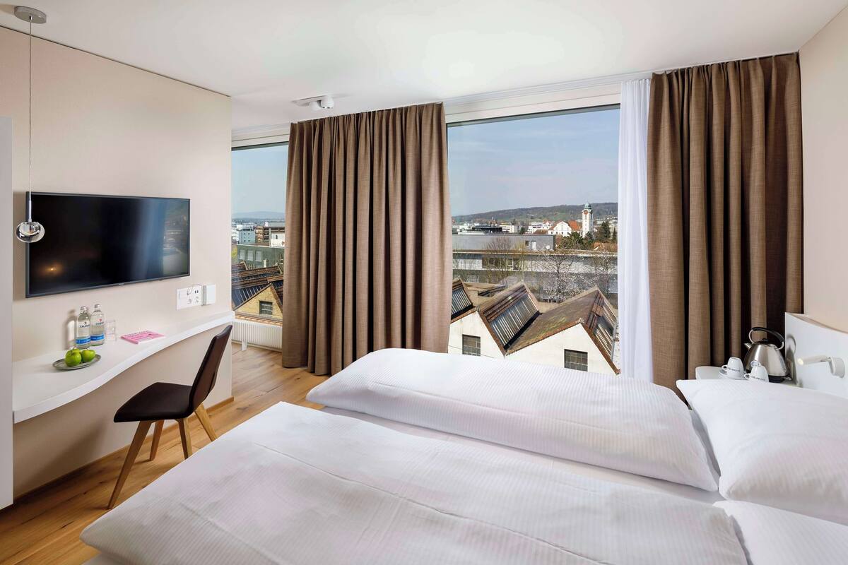 A hotel room with a bed, a desk with a TV, and a view of buildings.