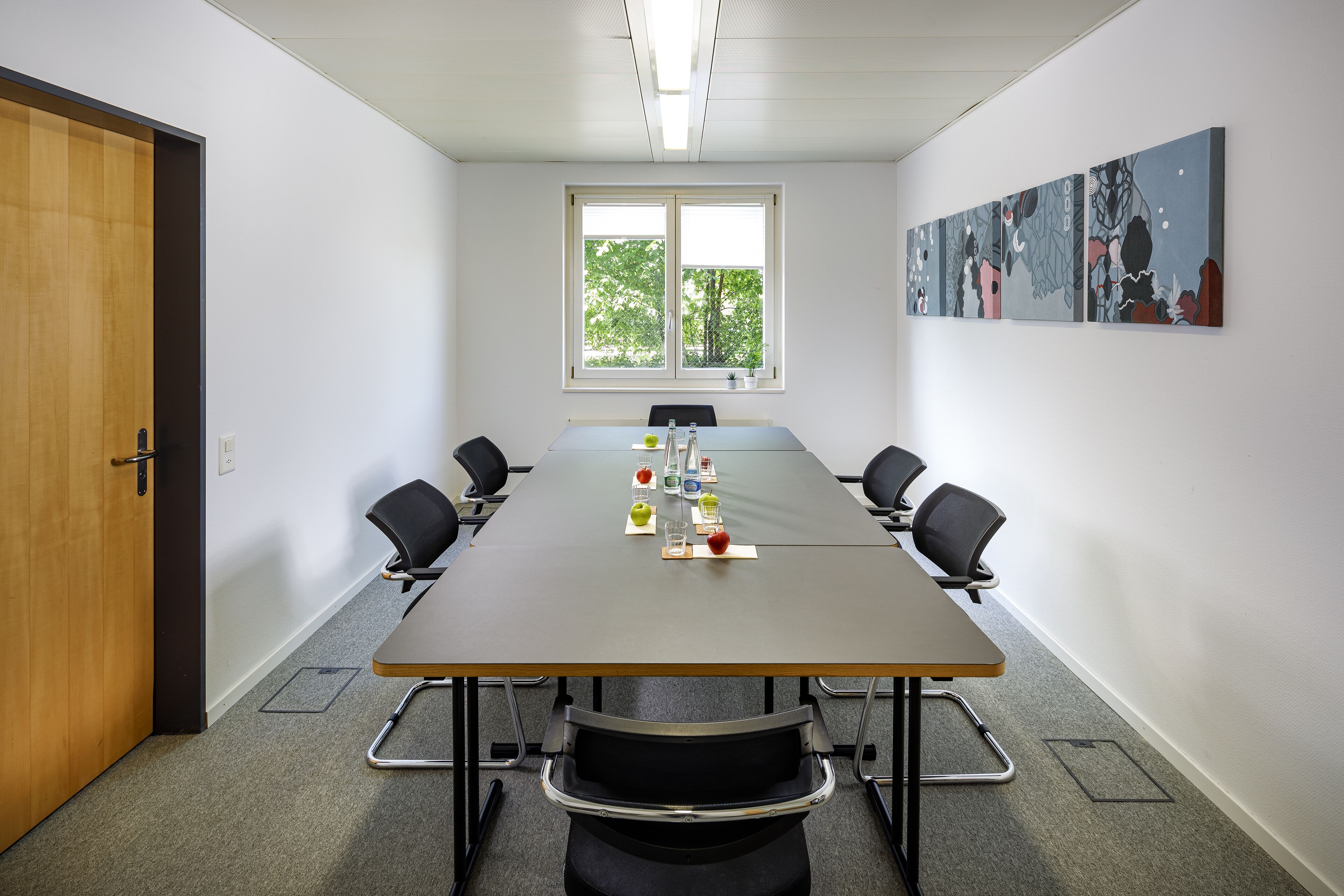 A conference room with a long table, four chairs, a window, and abstract art on the wall.