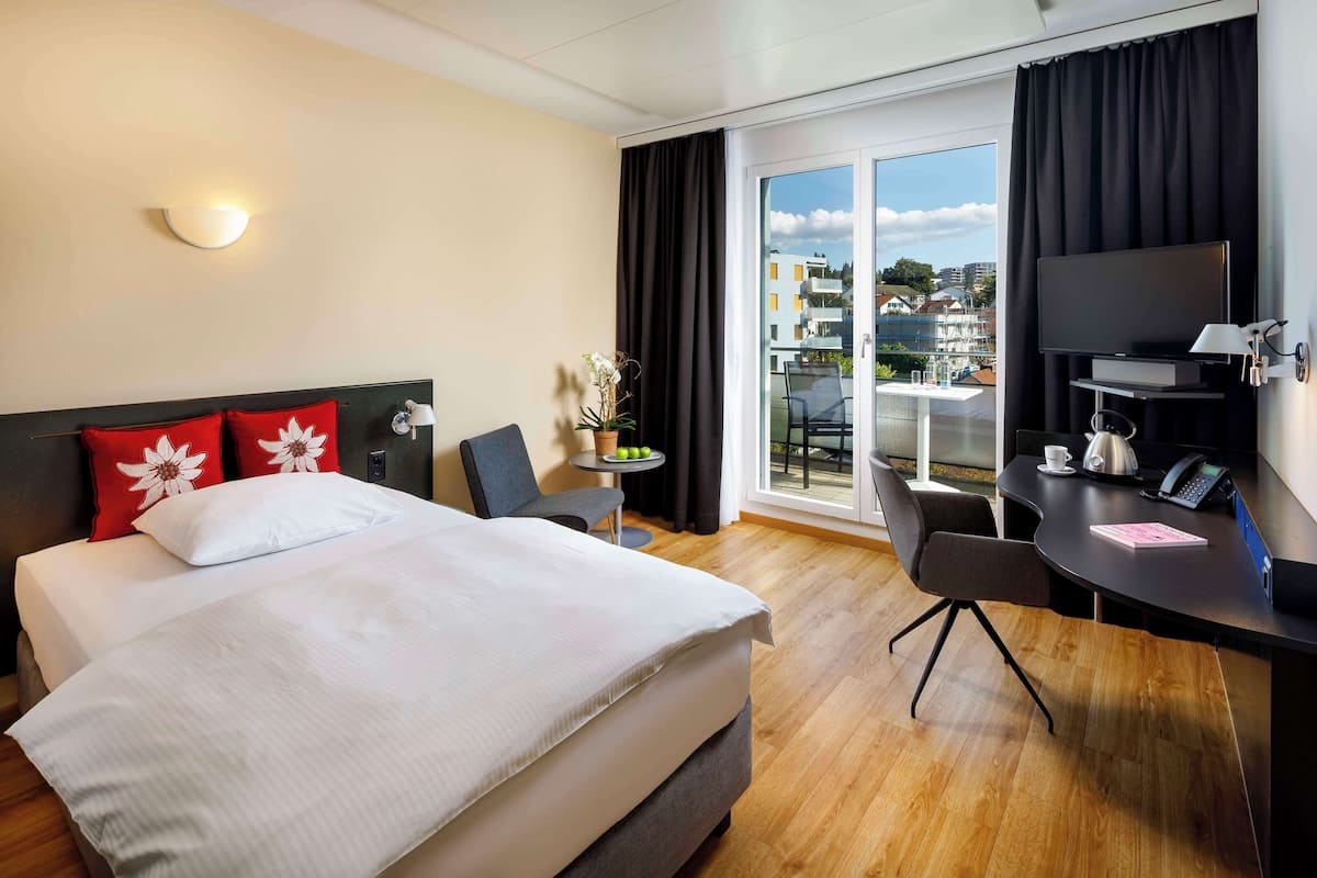 A hotel room with a bed, a desk, a chair, a TV, and a balcony with a city view.