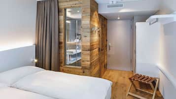 A hotel room with a bed, a wooden wardrobe, a small wooden stool, and a bathroom visible through a glass door.