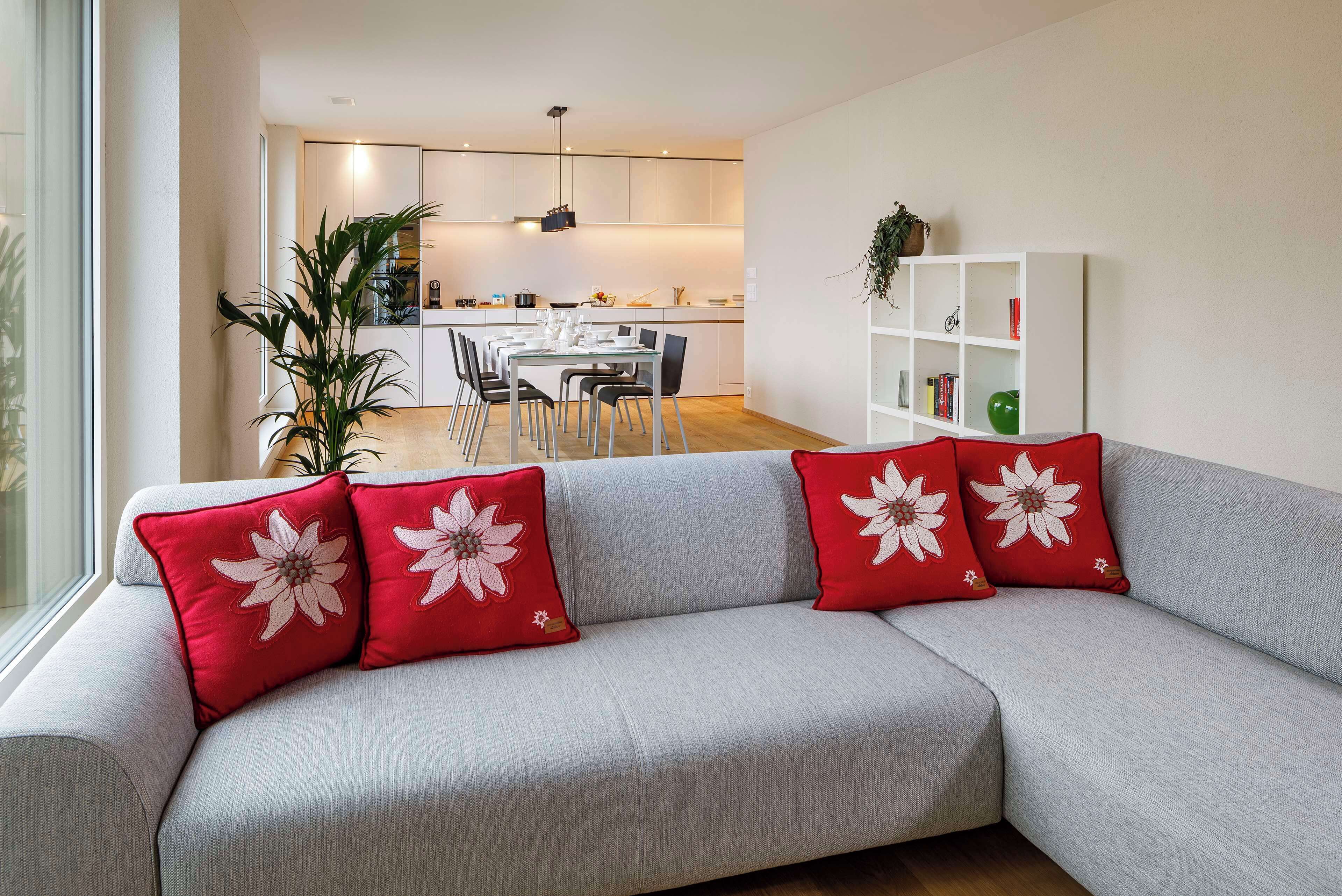 A modern living room with a grey sofa, red pillows with white floral designs, a white bookshelf, and a dining area in the background.