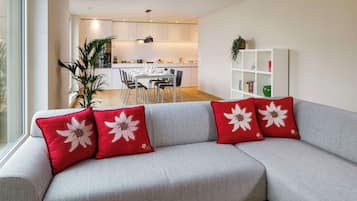 A modern living room with a grey sofa, red pillows with white floral designs, a white bookshelf, and a dining area in the background.