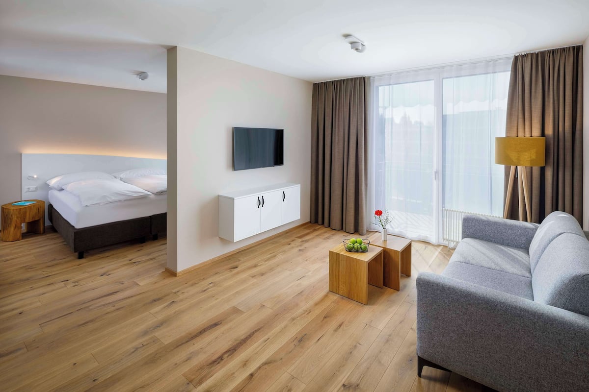 A modern hotel room with a bed, a sofa, a TV, and a small table with fruit.