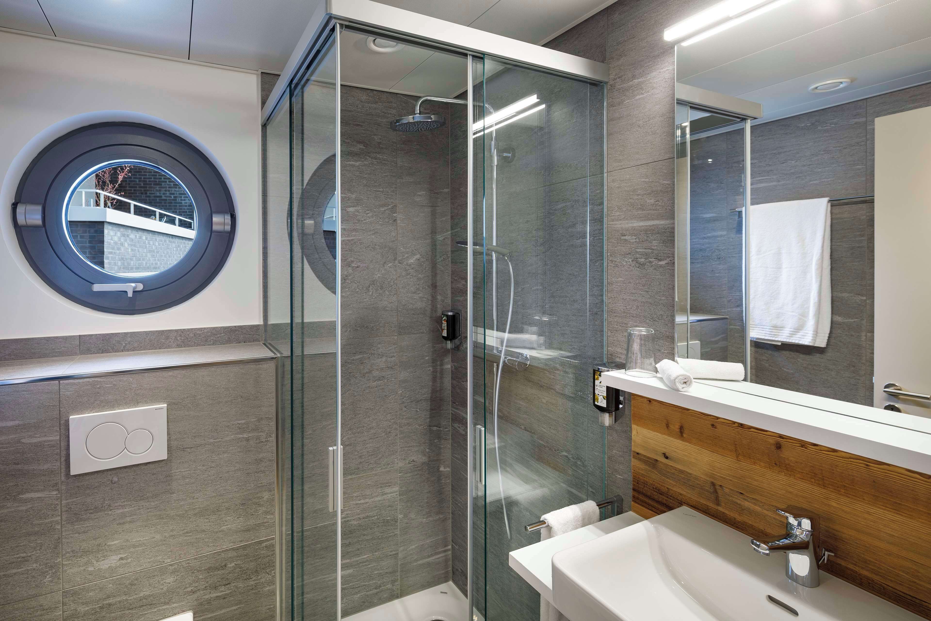 Een moderne badkamer met een douchecabine van glas, een rond raam en een houten wastafelmeubel.