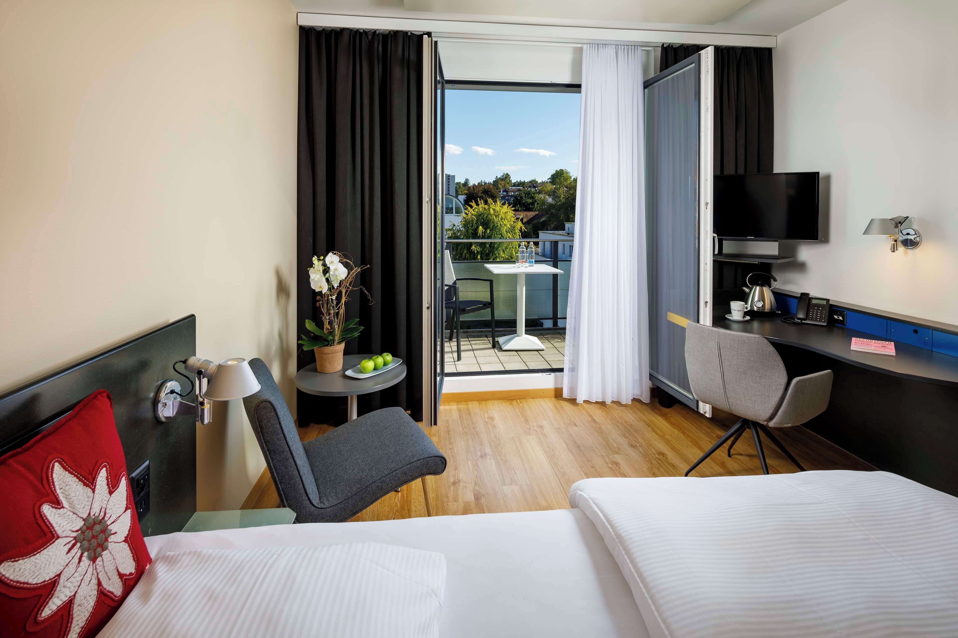 A hotel room with a bed, a desk, a chair, a TV, and a balcony with a table and chairs.
