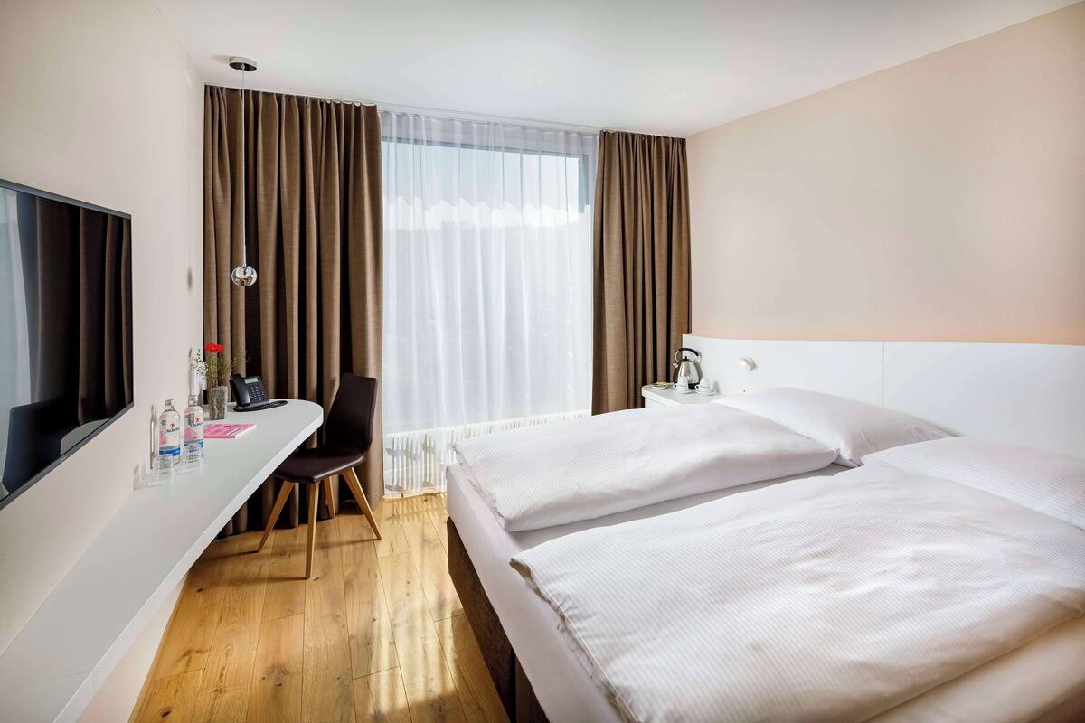A hotel room with a bed, a desk, a chair, a TV, and a window with curtains.