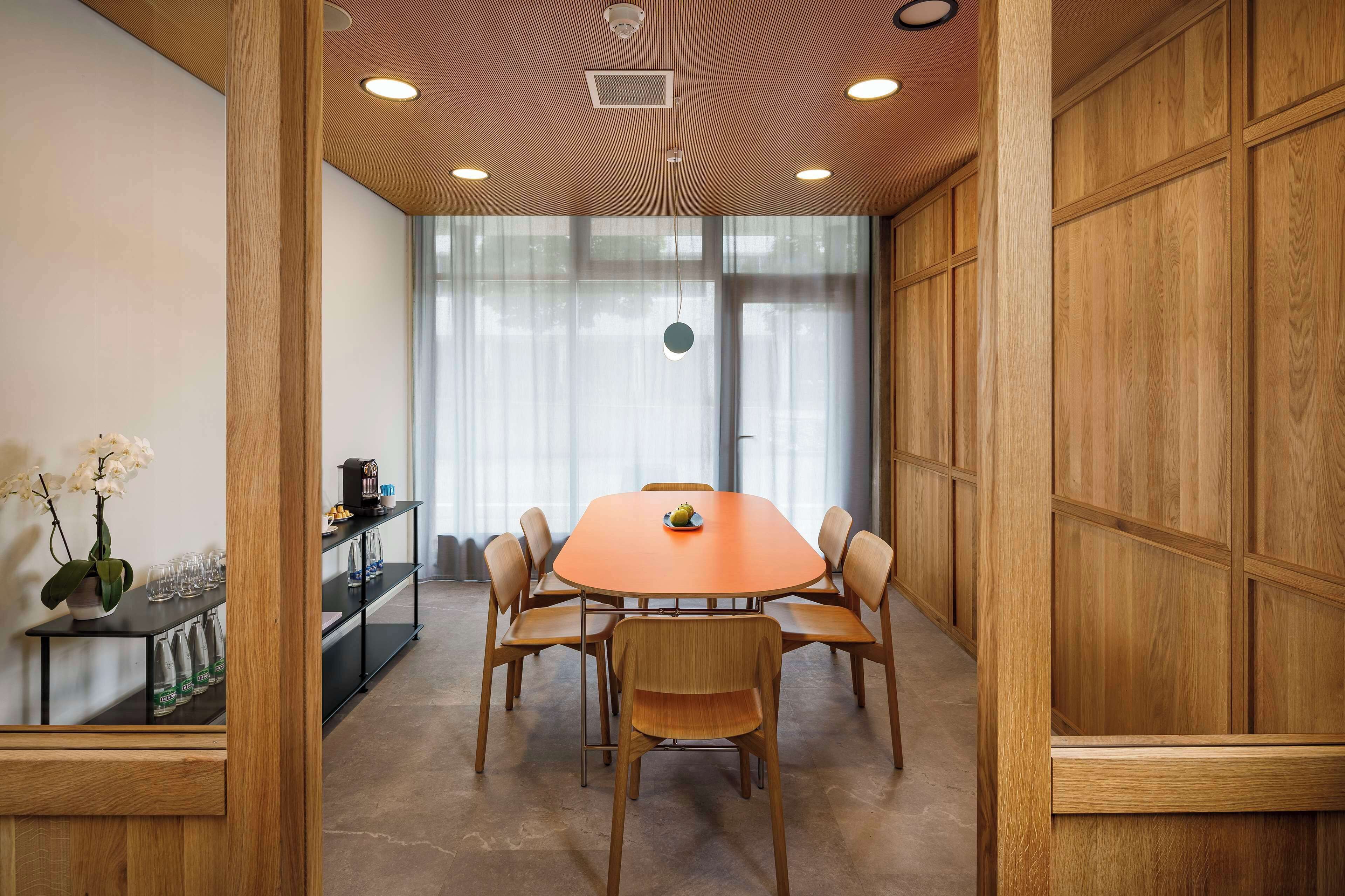 A conference room with a round table, six chairs, a vase with flowers, and a shelf with glasses and a coffee maker.