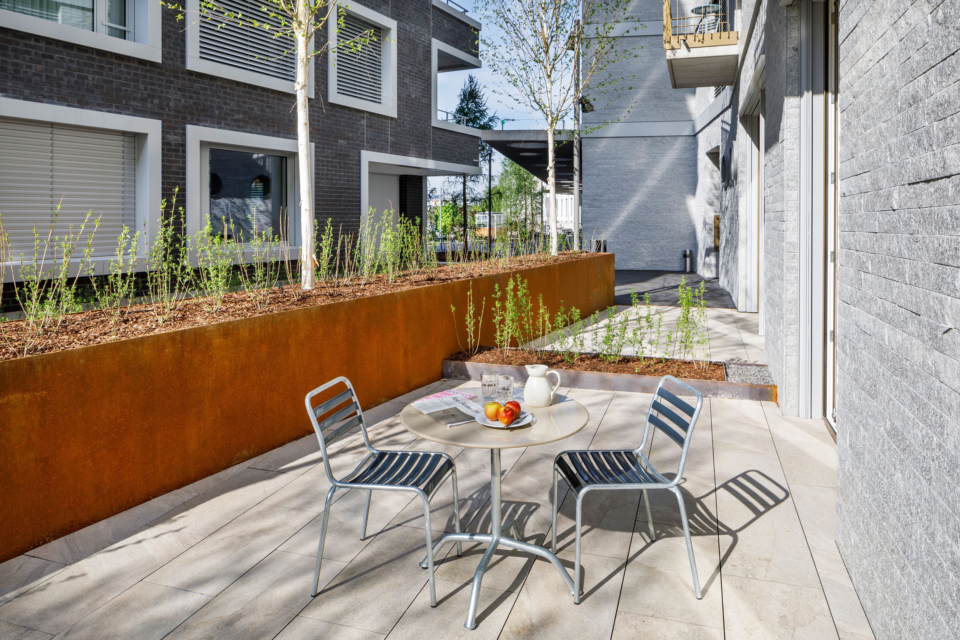 Eine Terrasse mit zwei Metallstühlen und einem Tisch, einer weißen Kanne und einem Teller mit Obst.
