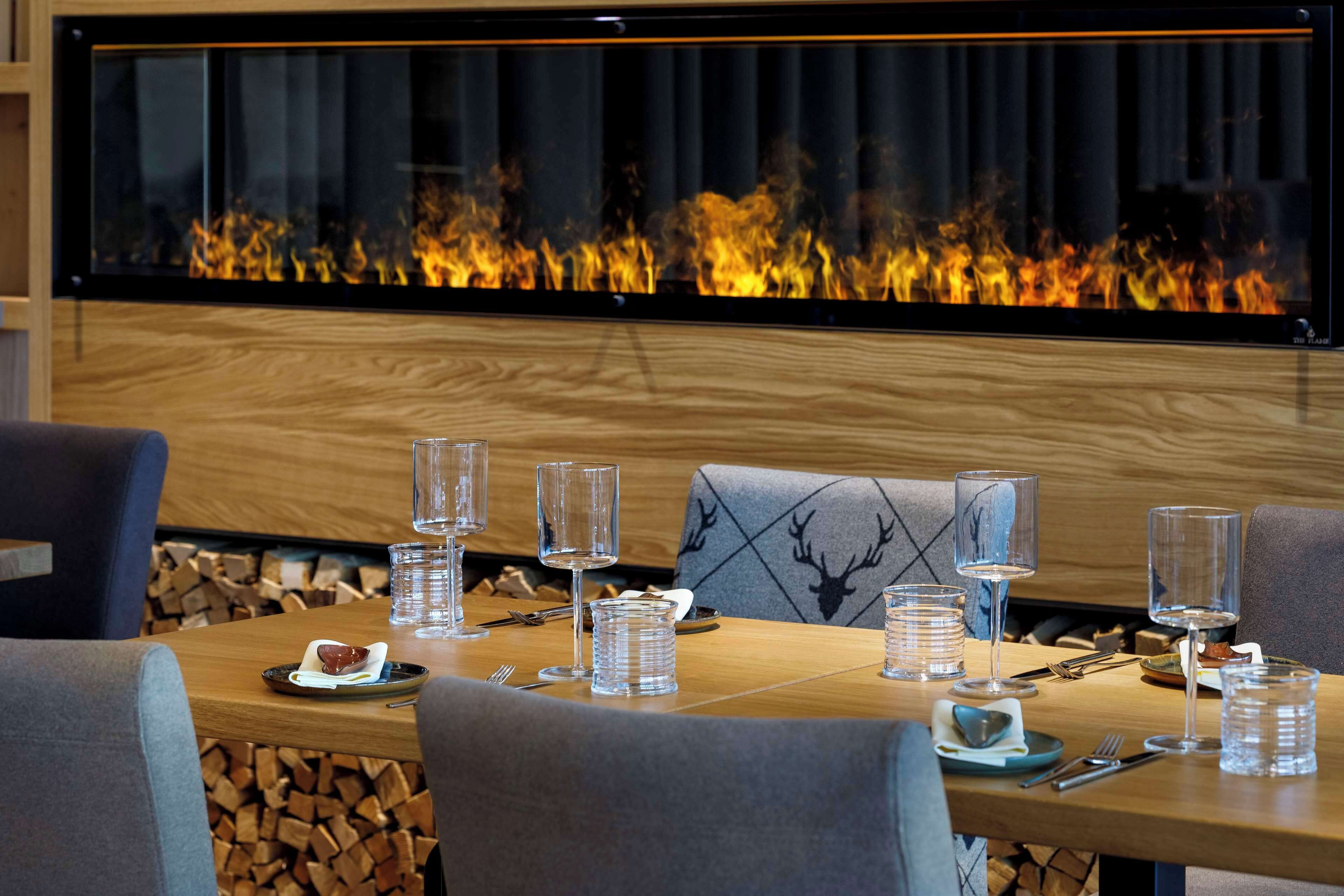 Un comedor con una mesa larga dispuesta para dos personas, una chimenea encendida y una pared de madera detrás de la mesa.