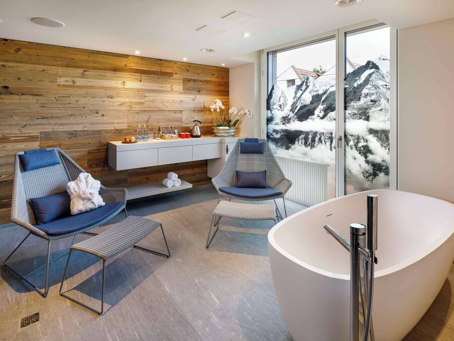 A modern bathroom with a large bathtub, two seating areas, a wooden wall, and a window with a mountain view.