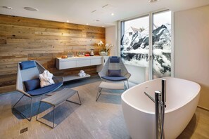 A modern bathroom with a large bathtub, two seating areas, a wooden wall, and a window with a mountain view.