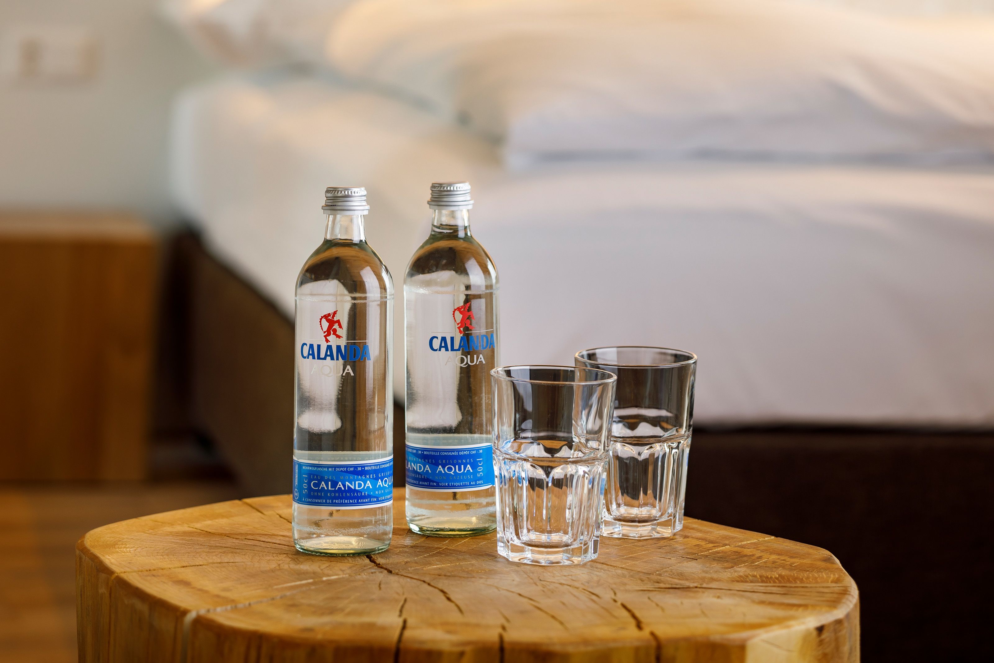 Two bottles of Calanda Aqua water and three glasses on a wooden table in a hotel room.