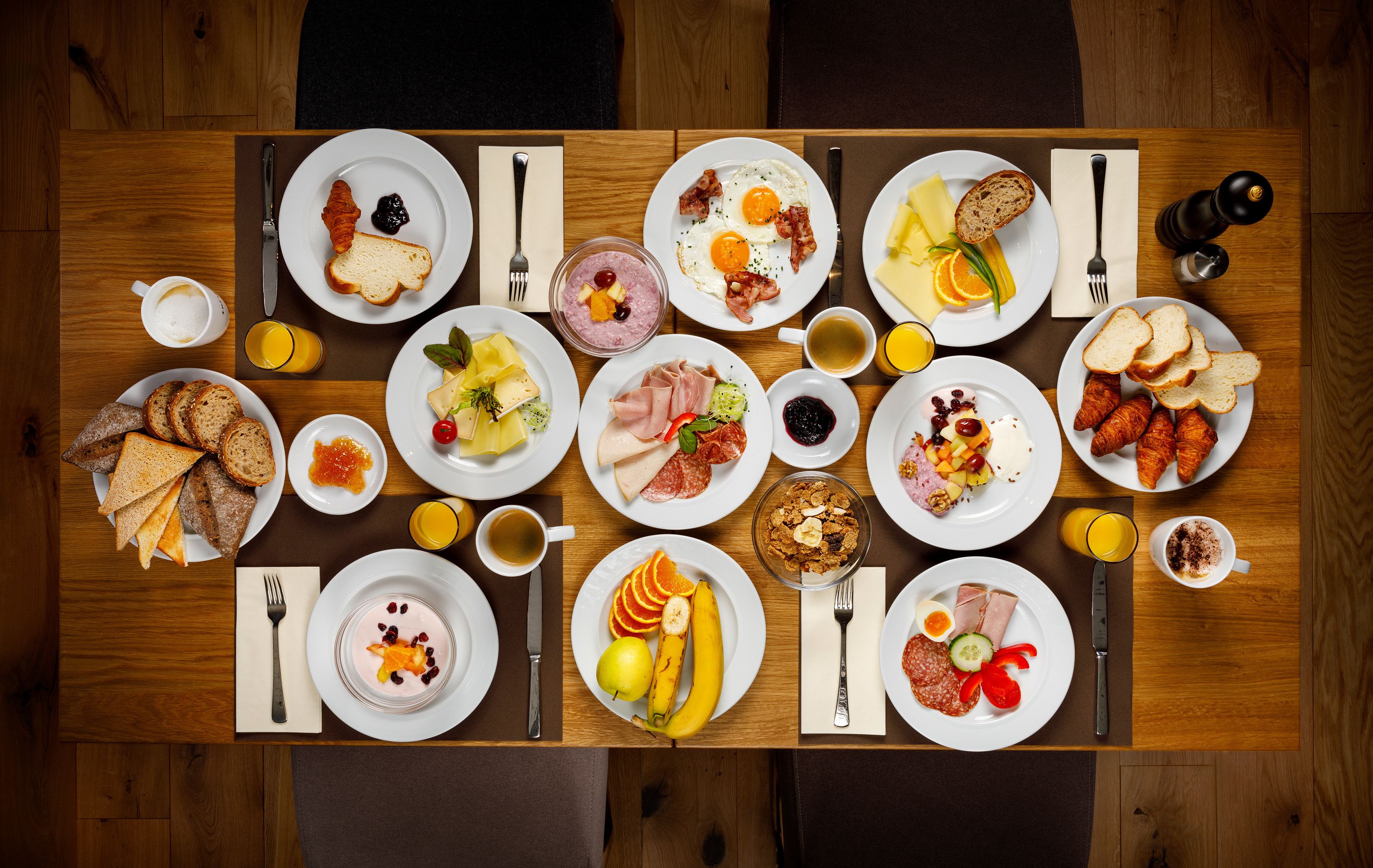 A wooden dining table with various breakfast items including bread, eggs, fruits, and beverages.