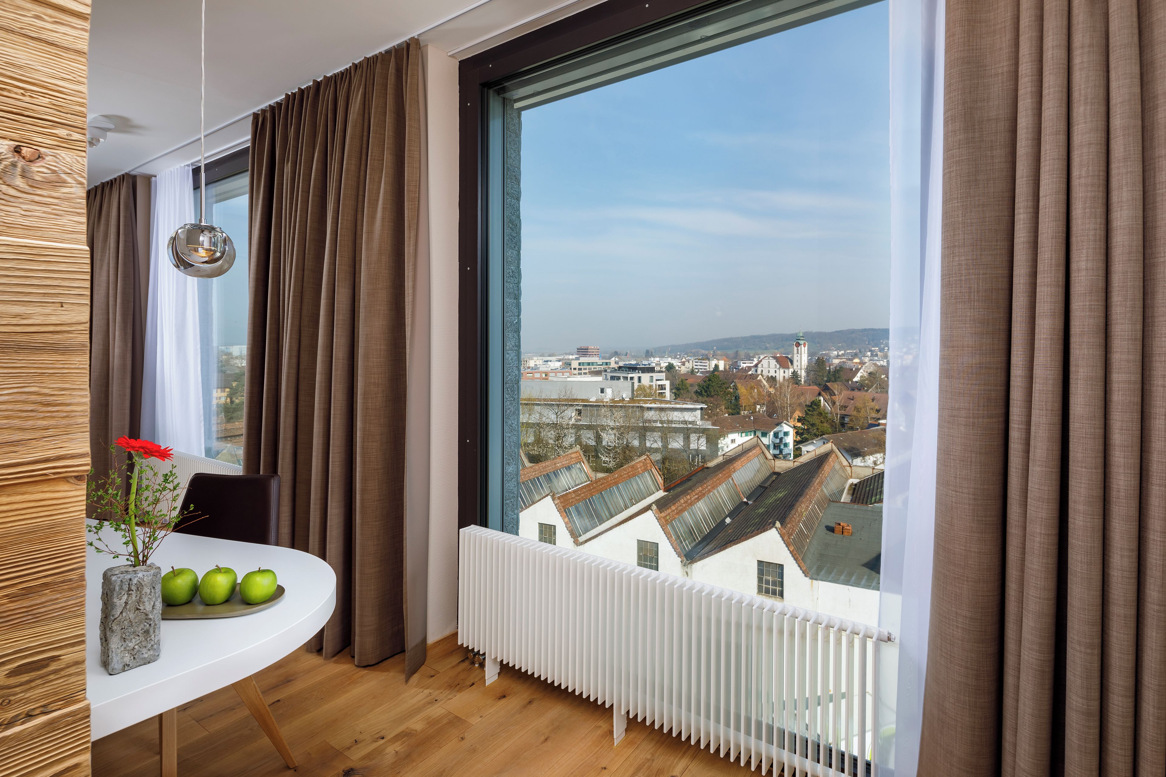 Ein Zimmer mit einem großen Fenster, einem Tisch mit Äpfeln und einer Vase, und Blick auf eine Stadtlandschaft.