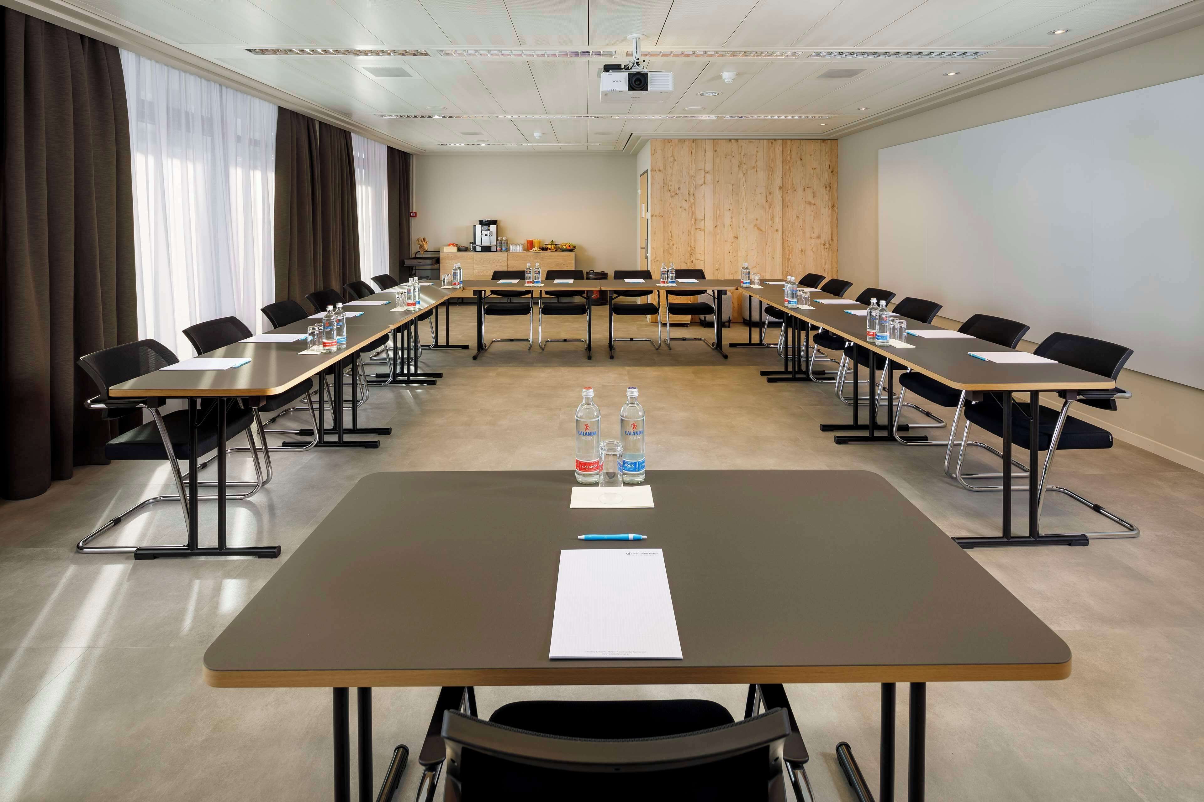 A conference room with long tables, chairs, water bottles, and a projector screen.