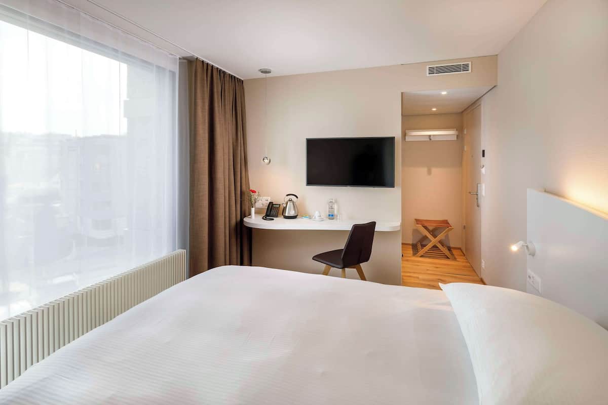 A hotel room with a large bed, a desk with a flat-screen TV, a chair, and a view of the city through the window.