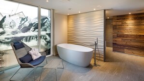 A modern bathroom with a freestanding bathtub, a chair with cushions, and a view of snow-capped mountains through large windows. - Hotel Allegra Lodge (Kloten)