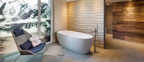 A modern bathroom with a freestanding bathtub, a chair with cushions, and a view of snow-capped mountains through large windows.