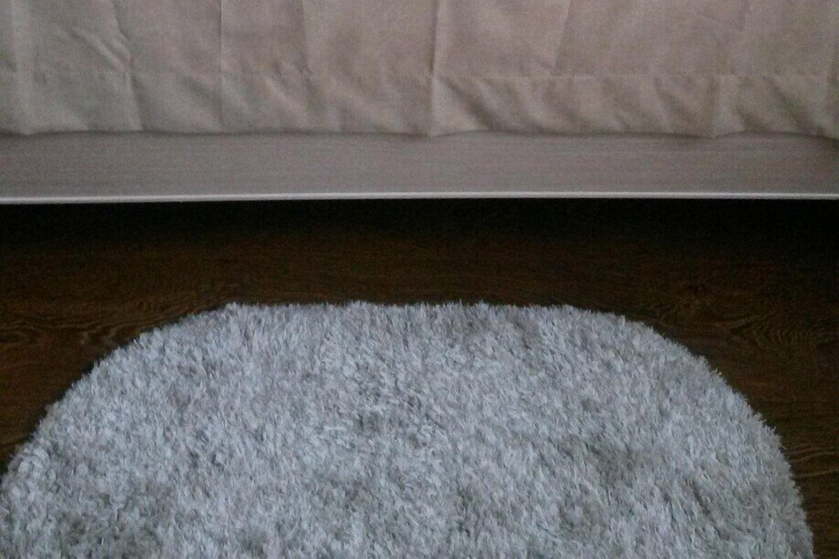 A neatly made bed with a beige bedspread and pillows, a framed picture on the wall, and a fluffy rug on the floor.