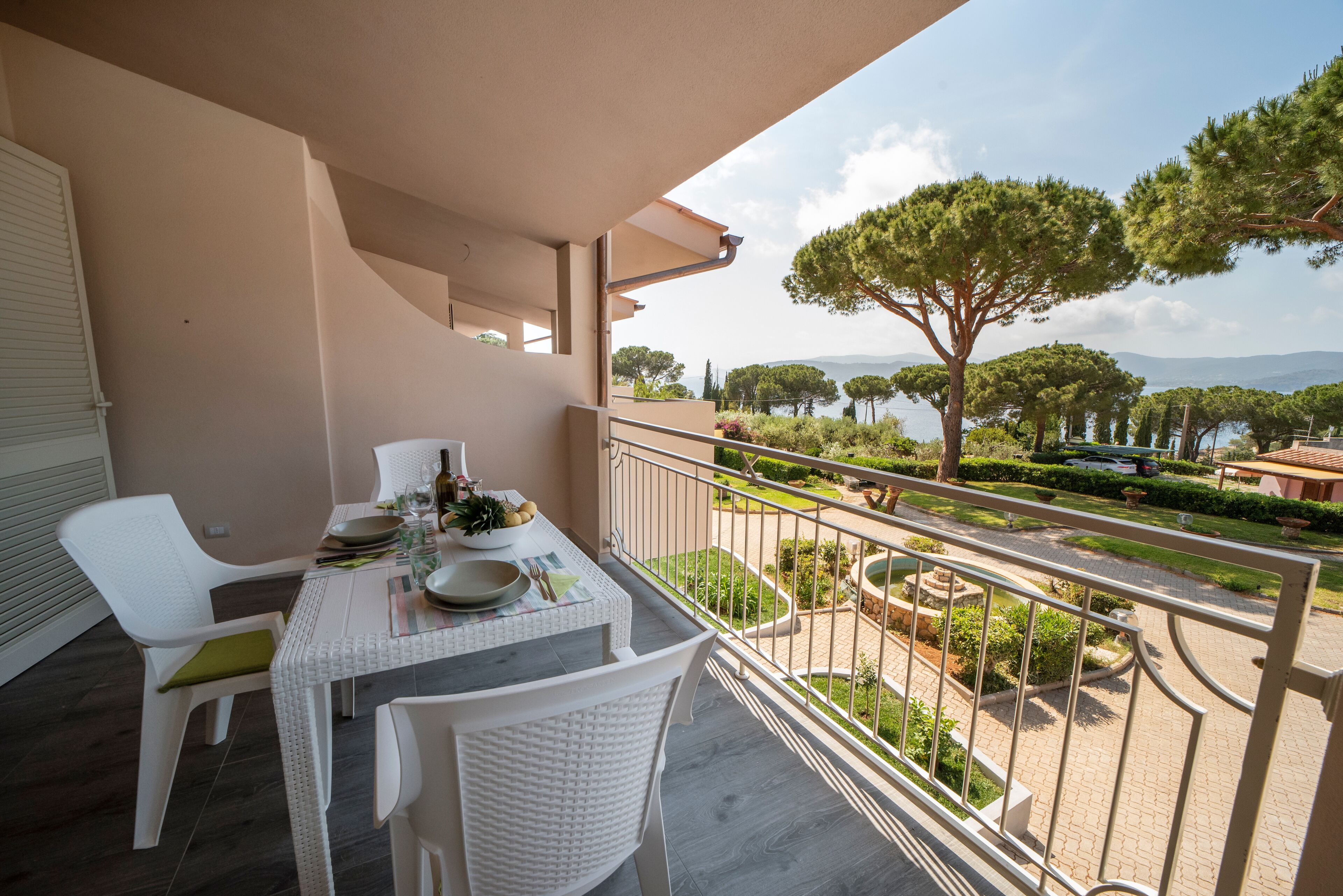 A balcony with a table set for dining, overlooking a landscaped garden and a body of water.