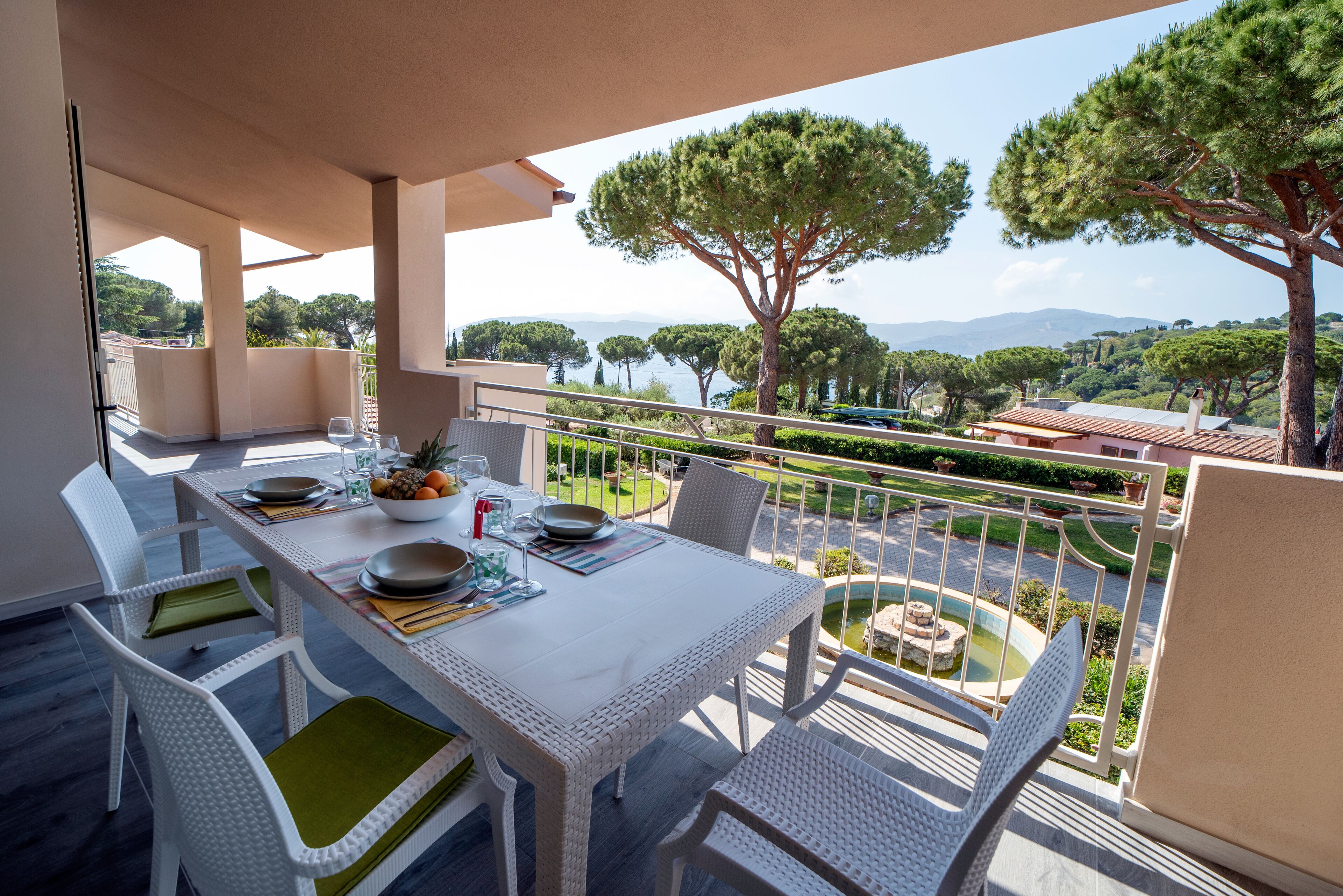 A balcony with a dining table set for four, overlooking a landscaped garden and a body of water.