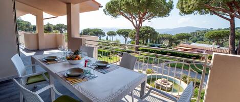 A balcony with a dining table set for four, overlooking a landscaped garden and a body of water.