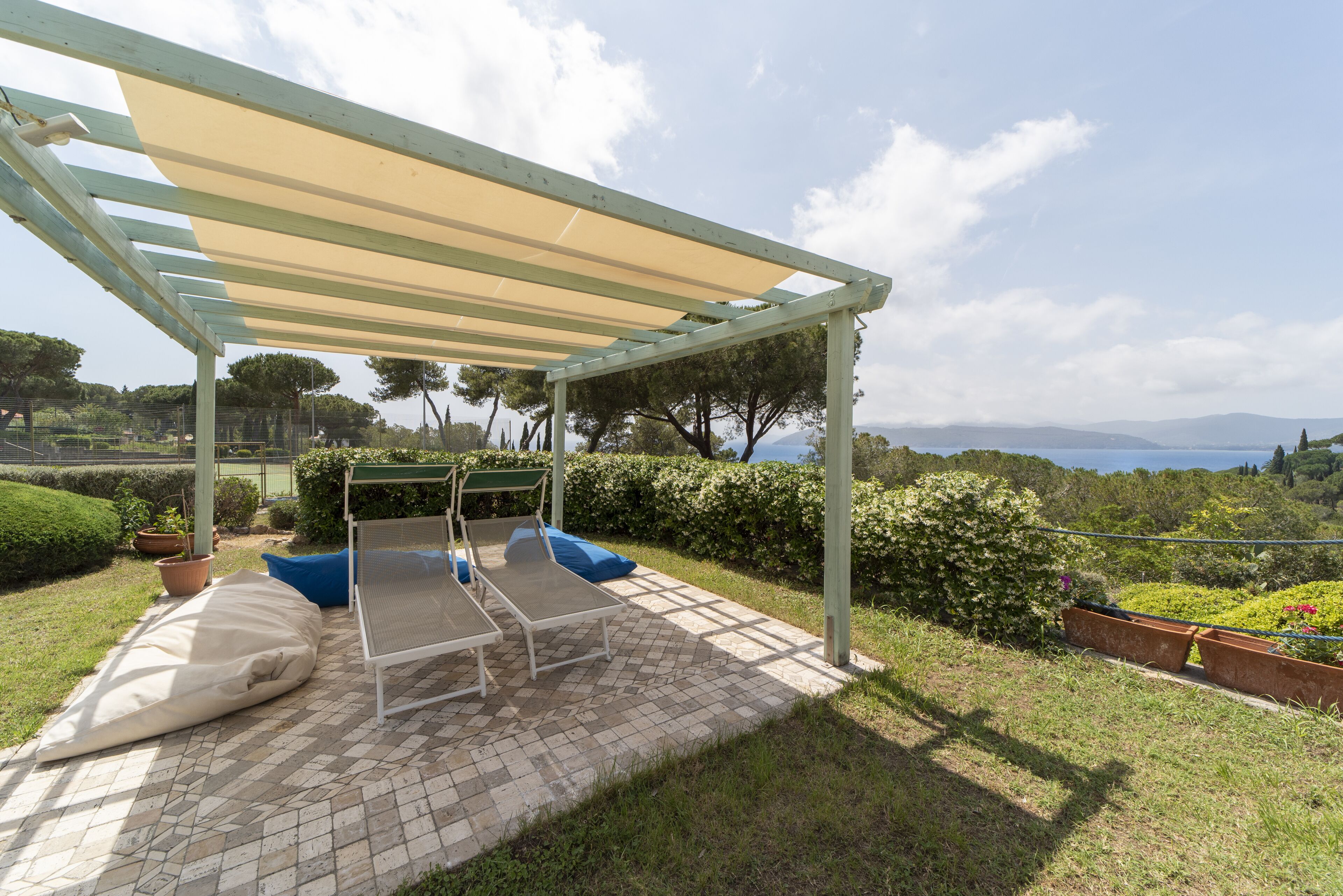 A covered outdoor seating area with a pergola, cushioned chairs, and a view of the landscape.