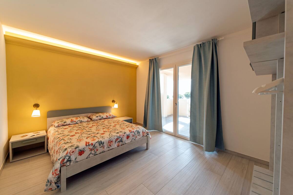 Une chambre à coucher avec un lit, des tables de chevet, une lampe fixée au mur et une vue à travers une porte vitrée.