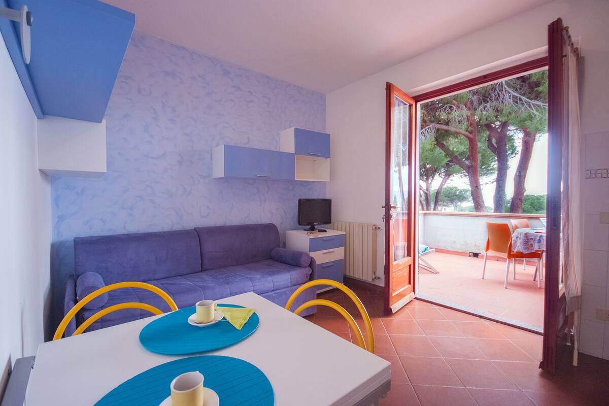 Une pièce avec un canapé bleu, une petite télévision, une table à manger avec des chaises jaunes et un balcon doté de mobilier d’extérieur.